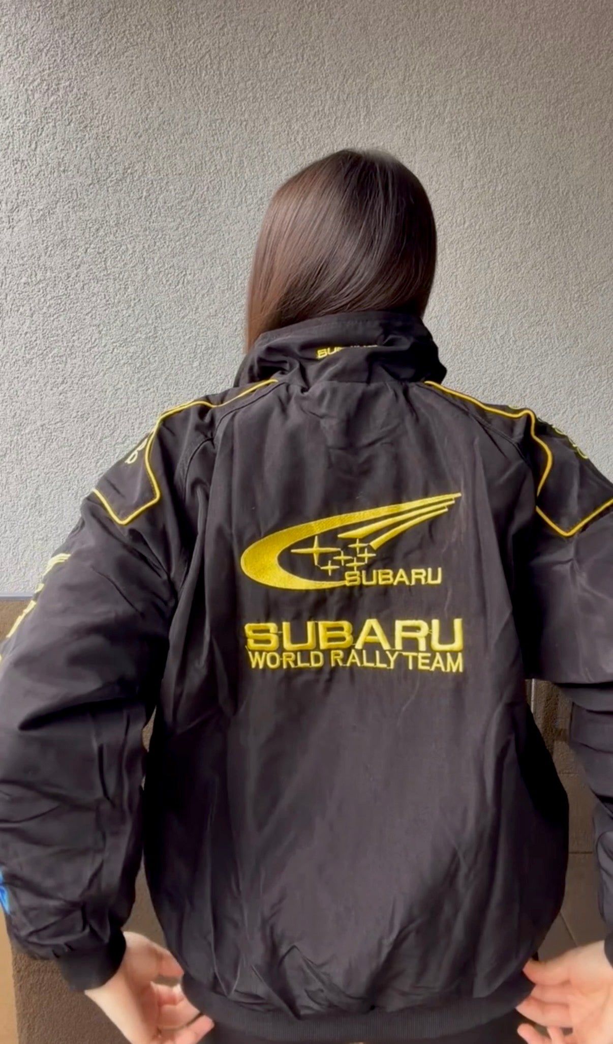 A person with long brown hair is seen from behind, wearing the Subaru Rally Vintage Racing Jacket featuring yellow accents and displaying the Subaru World Rally Team text along with the brand’s iconic logo on the back, highlighting Subaru’s rally heritage.