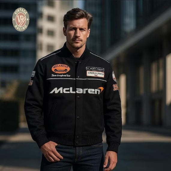 A man in a McLaren Vintage Bomber Jacket - Black stands outdoors in an urban setting, confidently facing the camera. The Vintage Racing logo is shown in the top left, reflecting motorsport-inspired streetwear style.