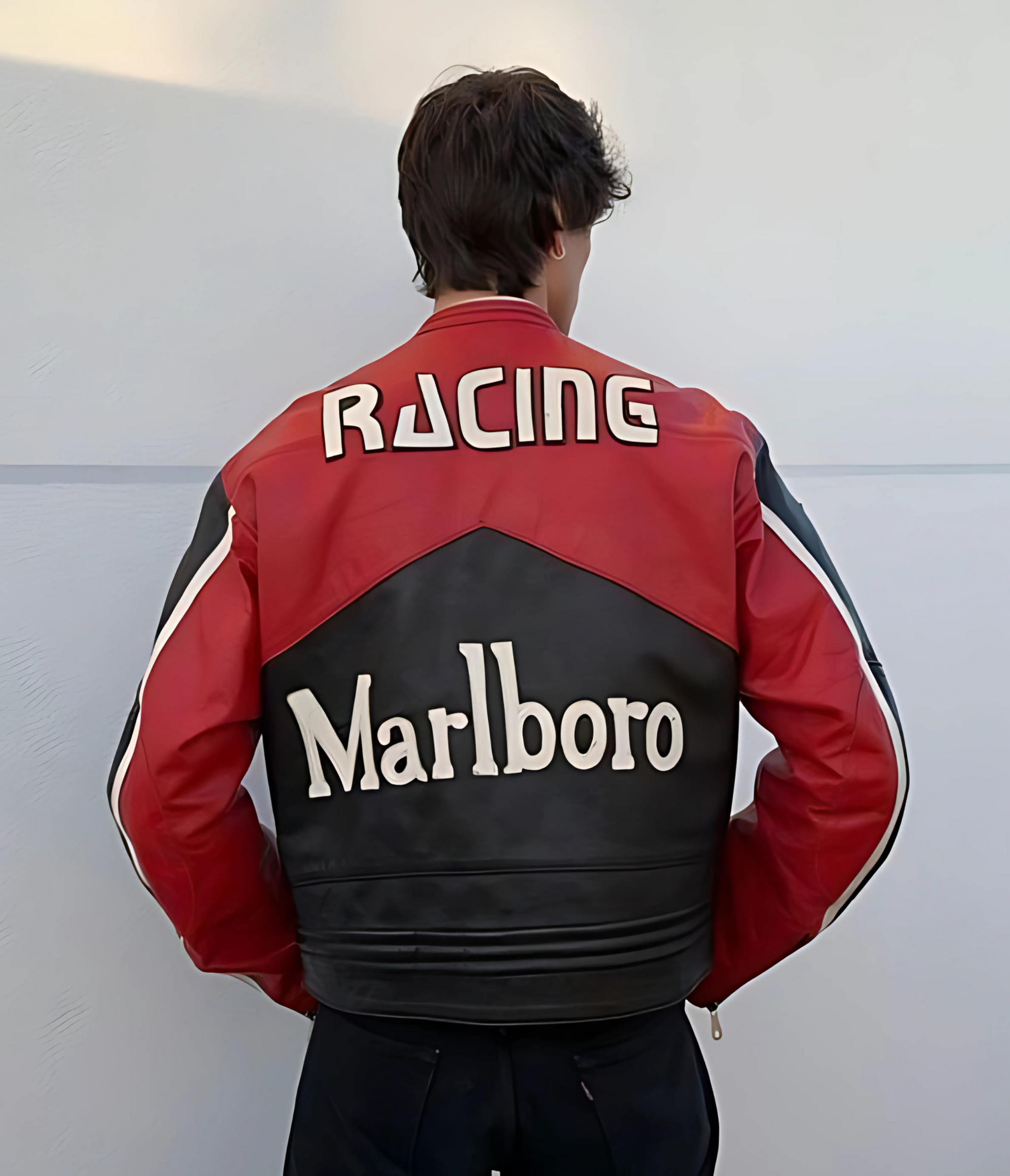 A person with dark hair faces away, wearing the Marlboro Racing Leather Jacket - Black Edition, a standout motorsport streetwear piece in black, against a plain light-colored wall.