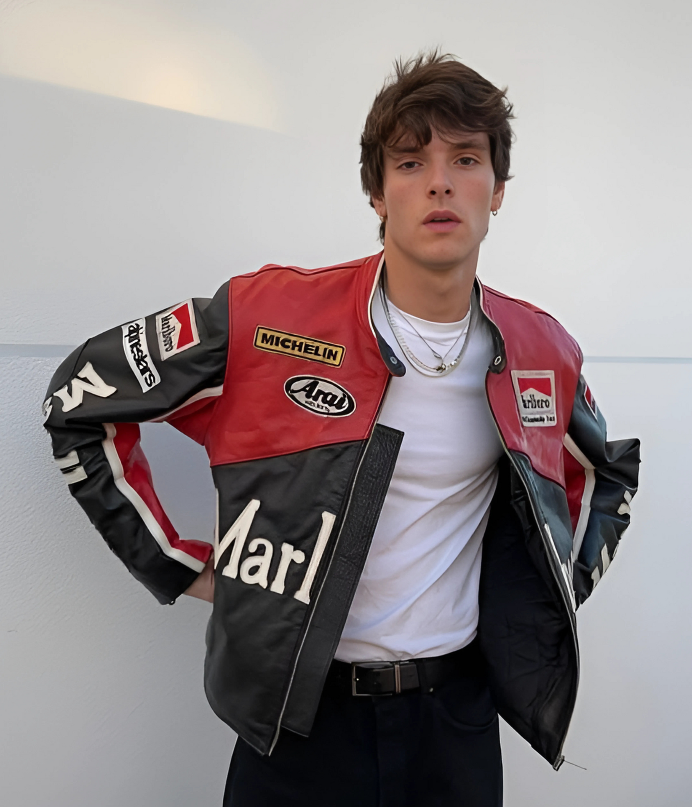 A young man wears the Marlboro Racing Leather Jacket - Black Edition with red, black, and white details and racing patches, standing against a light-colored wall and adjusting his motorsport streetwear.