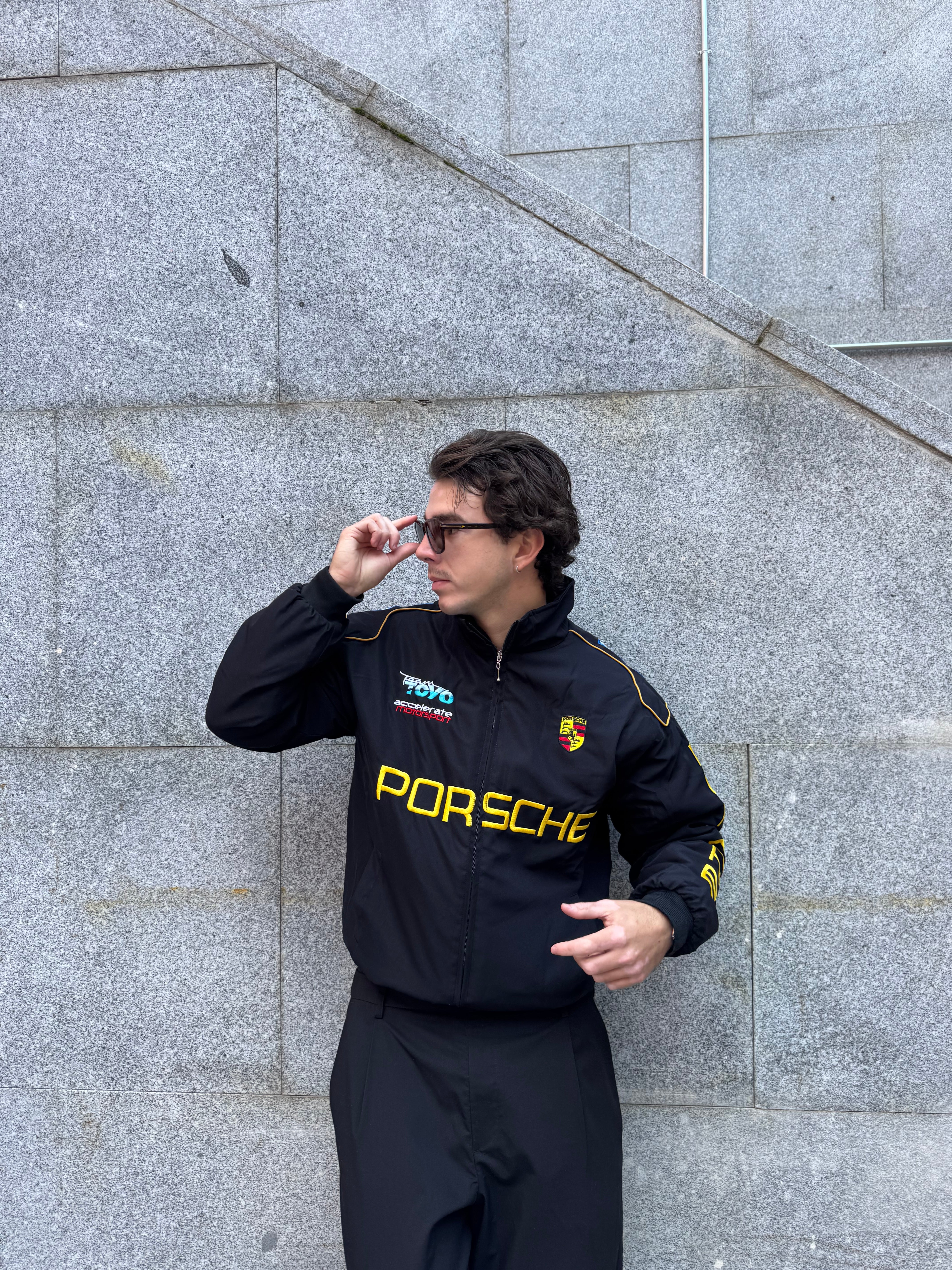 A man in sunglasses and a Porsche Vintage Racing Jacket stands against a gray stone wall, looking to the side as he adjusts his sunglasses.