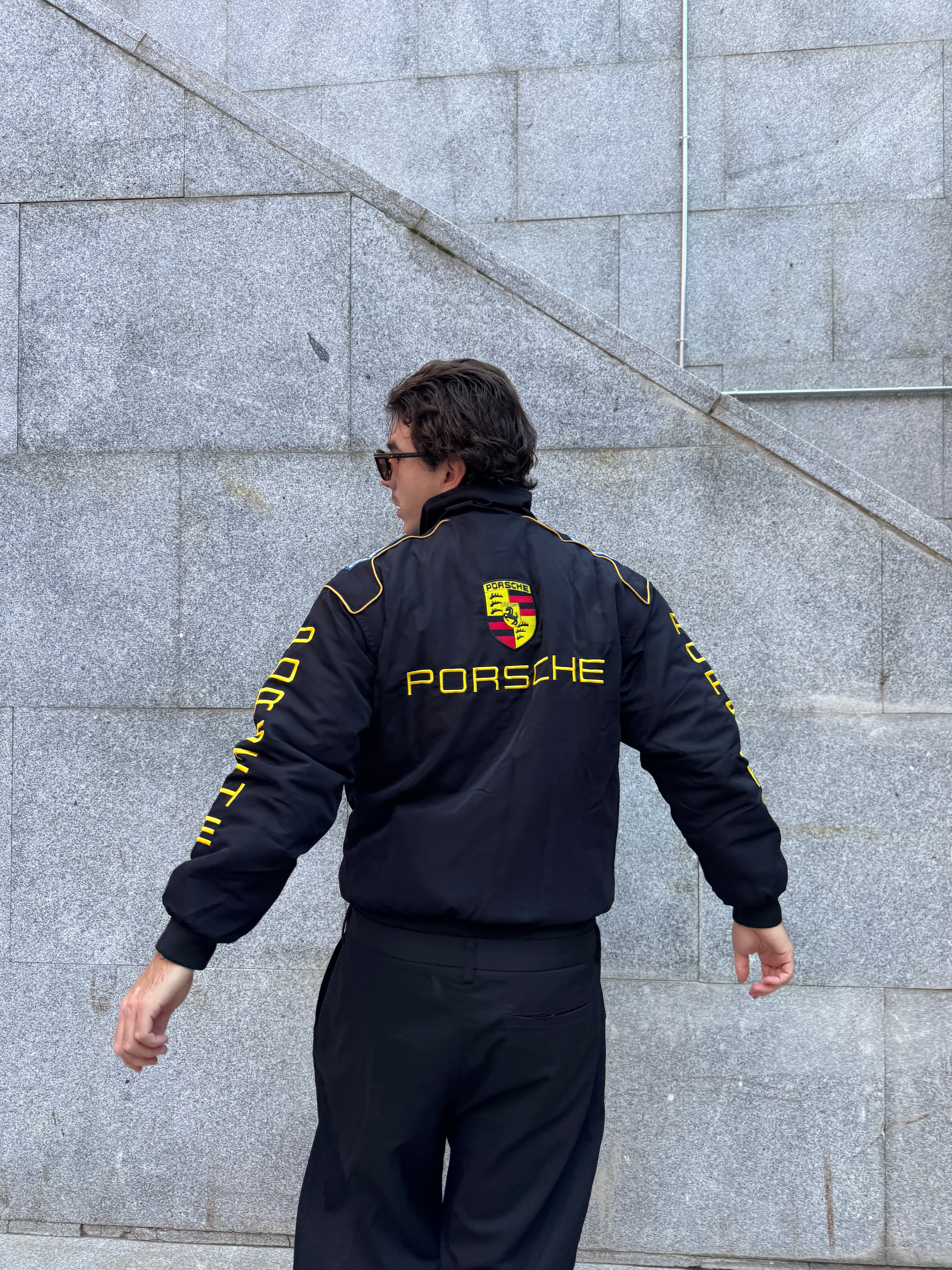 A person wearing the Porsche Vintage Racing Jacket, featuring black fabric with yellow lettering and logo, stands facing away from the camera in front of a gray stone wall, arms extended.