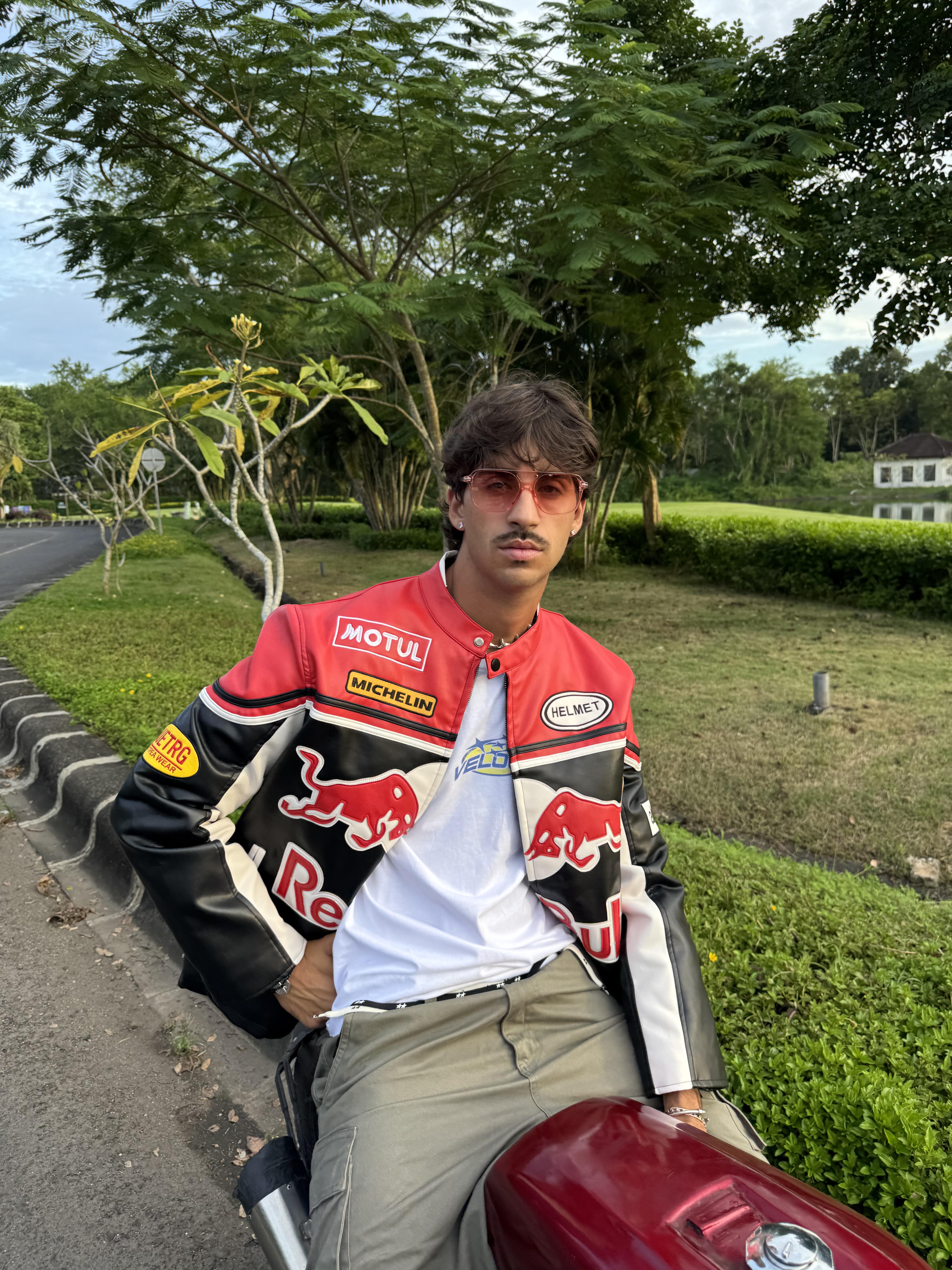 A man with brown hair, mustache, and sunglasses wears the Red Bull Leather Racing Jacket while sitting on a red motorcycle by a roadside surrounded by greenery and trees.