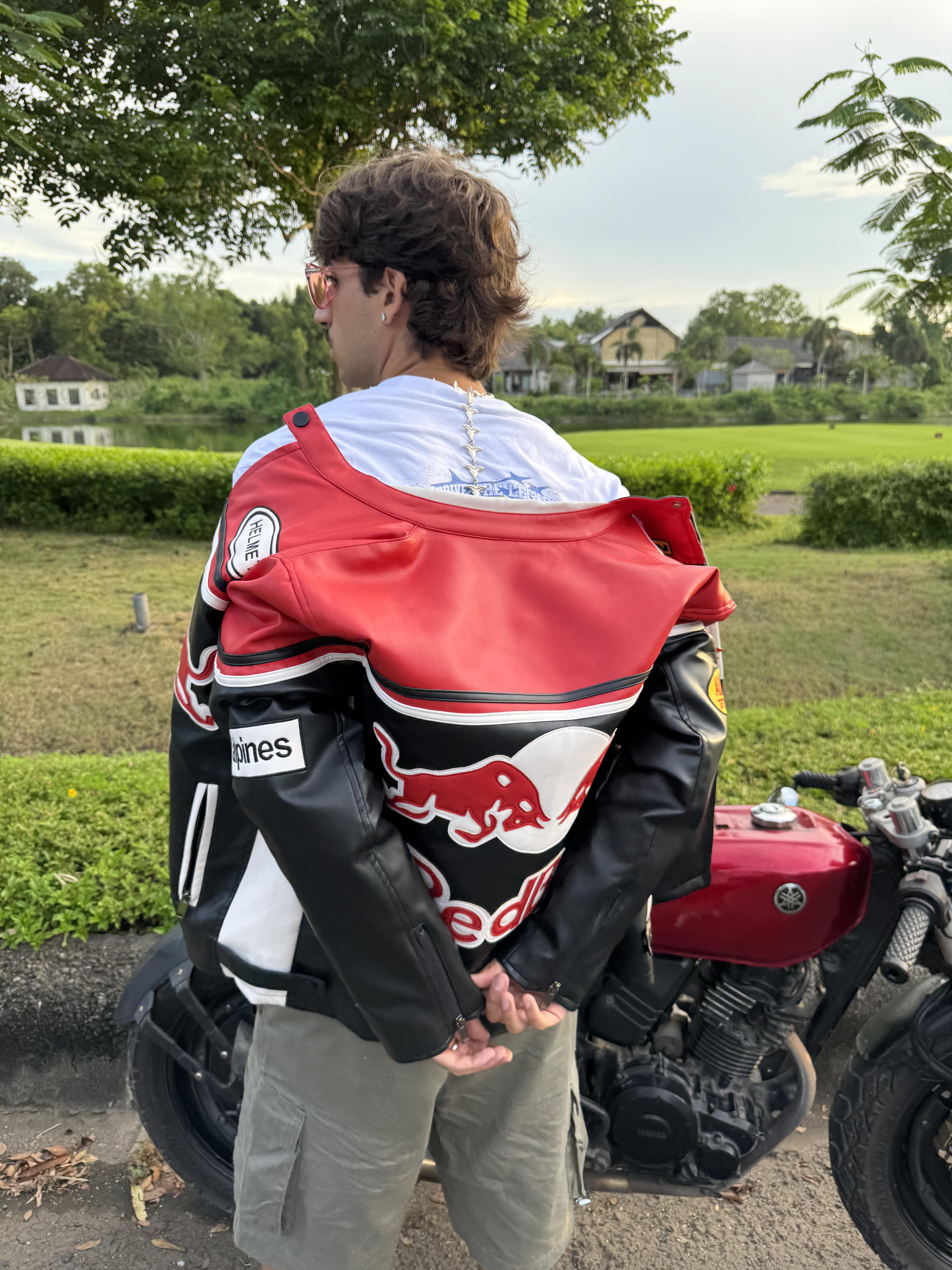 A person with brown hair, facing away in a white shirt, drapes the Red Bull Leather Racing Jacket over their shoulders. With hands clasped behind their back, they stand near a red motorcycle that echoes racing heritage.