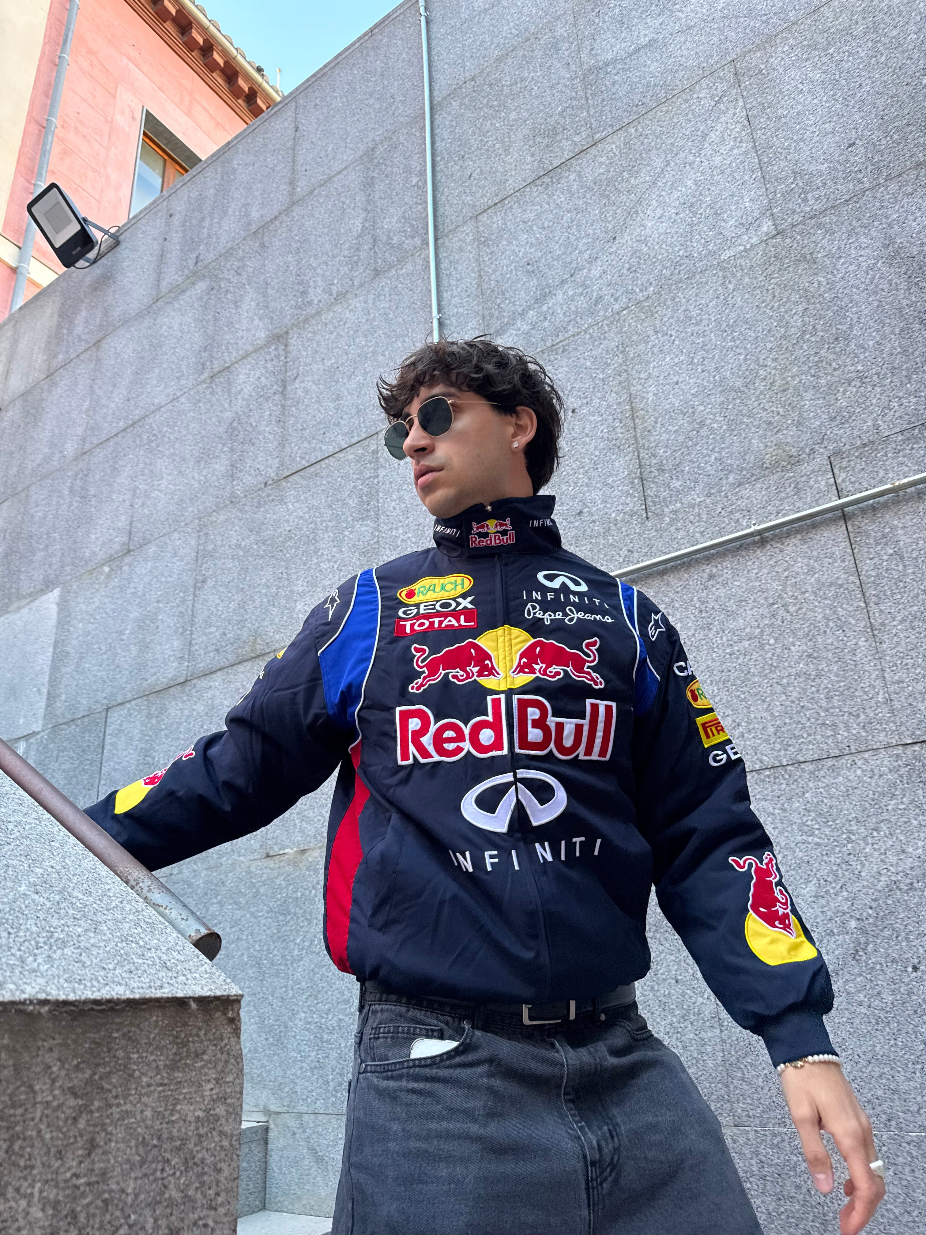 A man in sunglasses wears a Red Bull Racing F1 Jacket, standing confidently on outdoor steps next to a gray stone wall, gazing to the side.