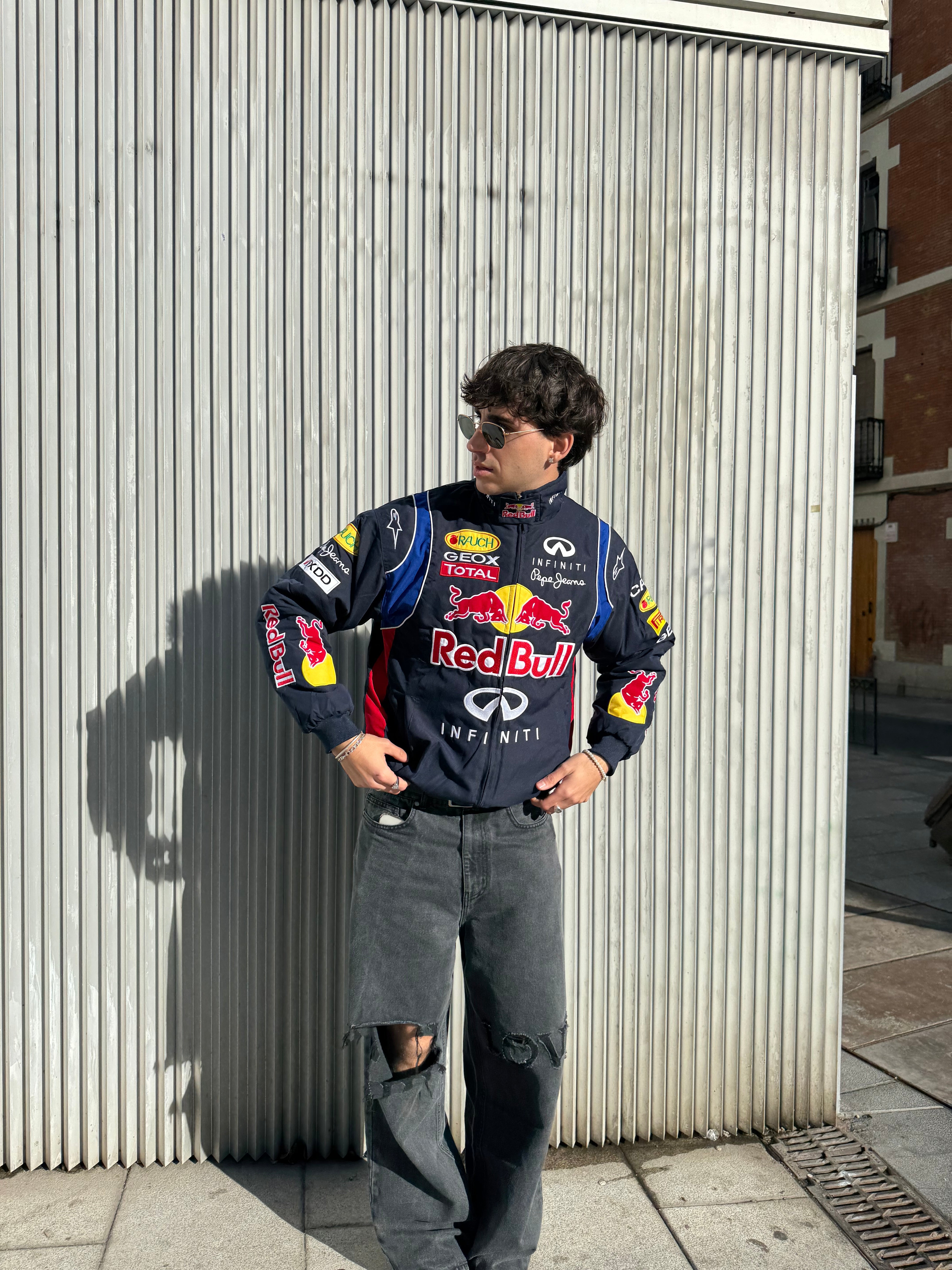A man wearing sunglasses and a Red Bull Racing F1 Jacket stands on a sunny sidewalk against a corrugated metal wall, hands on his hips, looking to the side.