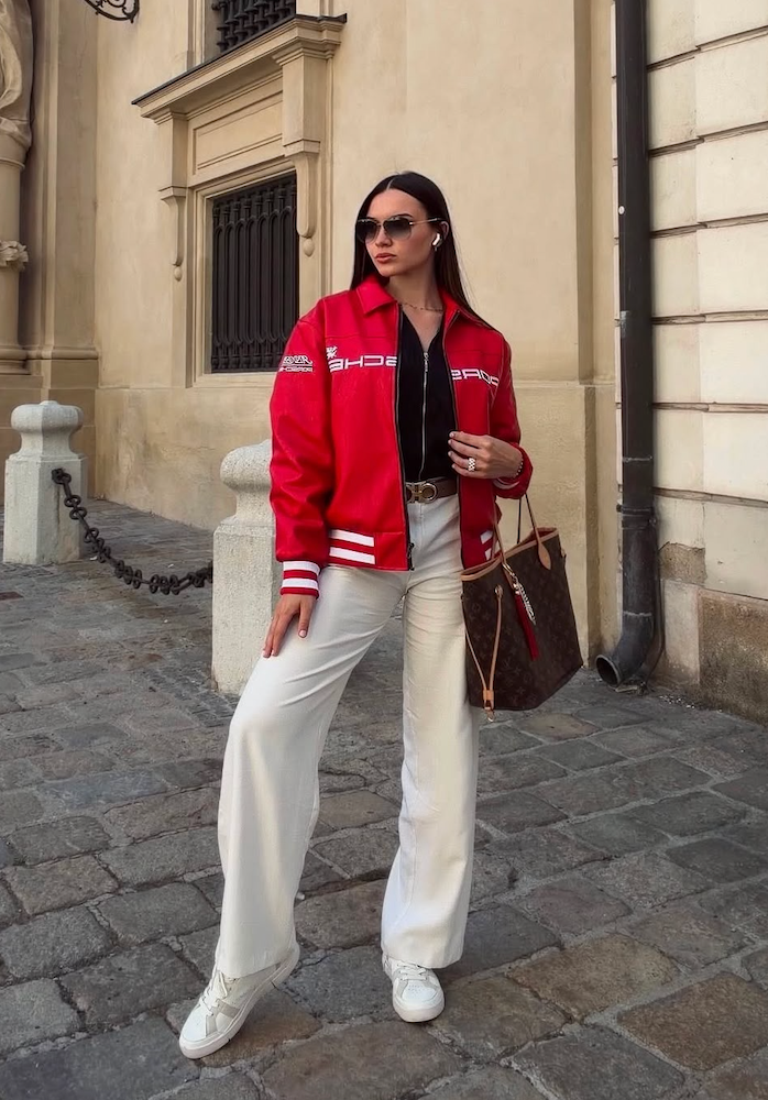 A woman wearing the Vintage Porsche Leather Jacket - Red Edition, paired with white pants and white sneakers, stands on a cobblestone street holding a brown handbag and wearing sunglasses.