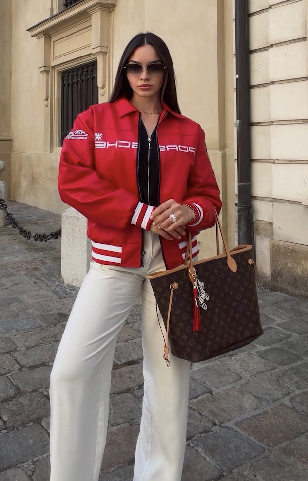 A woman poses on a cobblestone street wearing the Vintage Porsche Leather Jacket - Red Edition, paired with white pants and sunglasses, while holding a Louis Vuitton tote bag adorned with a scarf. She stands in front of a beige building.