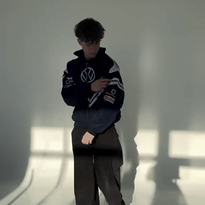 A young person with wavy hair stands in a sunlit room, wearing the Volkswagen Vintage Racing Jacket and loose brown pants, as bold geometric window shadows create a striking modern contrast behind them.