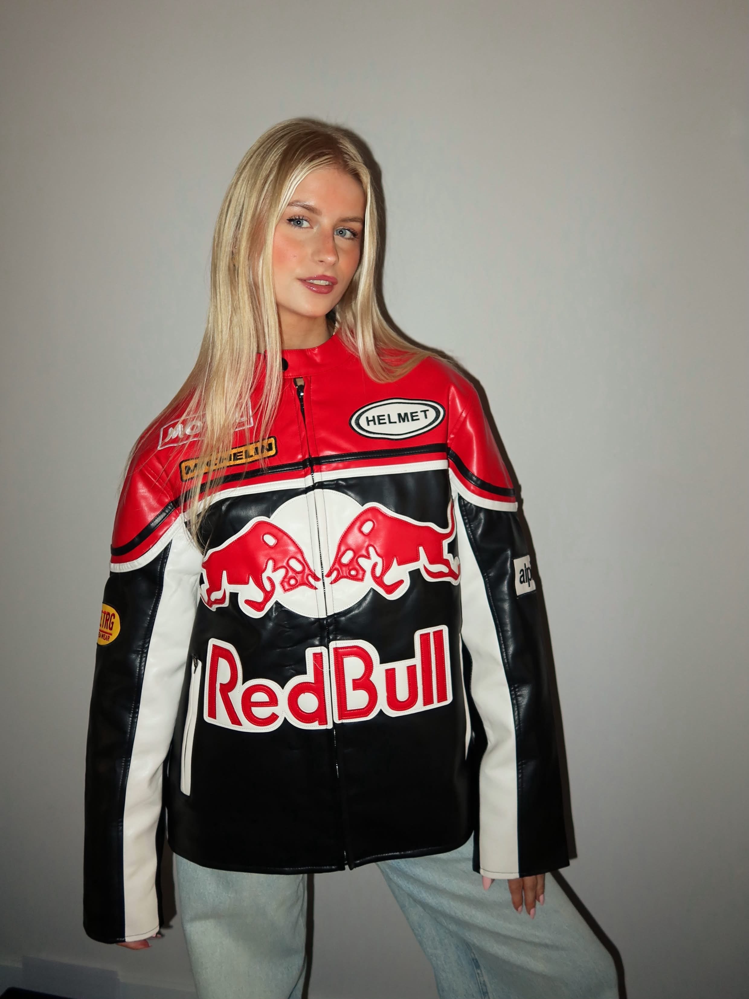 A woman with long blonde hair poses against a plain light-colored wall, wearing the Red Bull Leather Racing Jacket in black, red, and white with her arms slightly out.