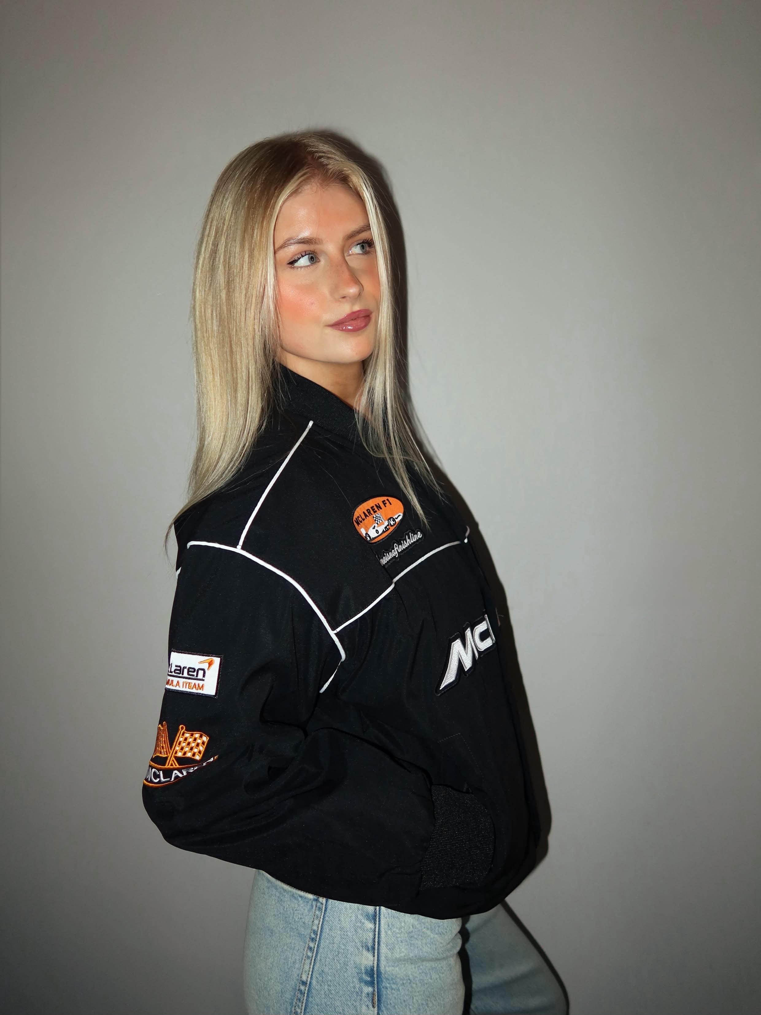 A woman with long blonde hair wears the McLaren Vintage Bomber Jacket in black, featuring orange patches, paired with light blue jeans. She stands against a plain gray background, looking slightly to the side.