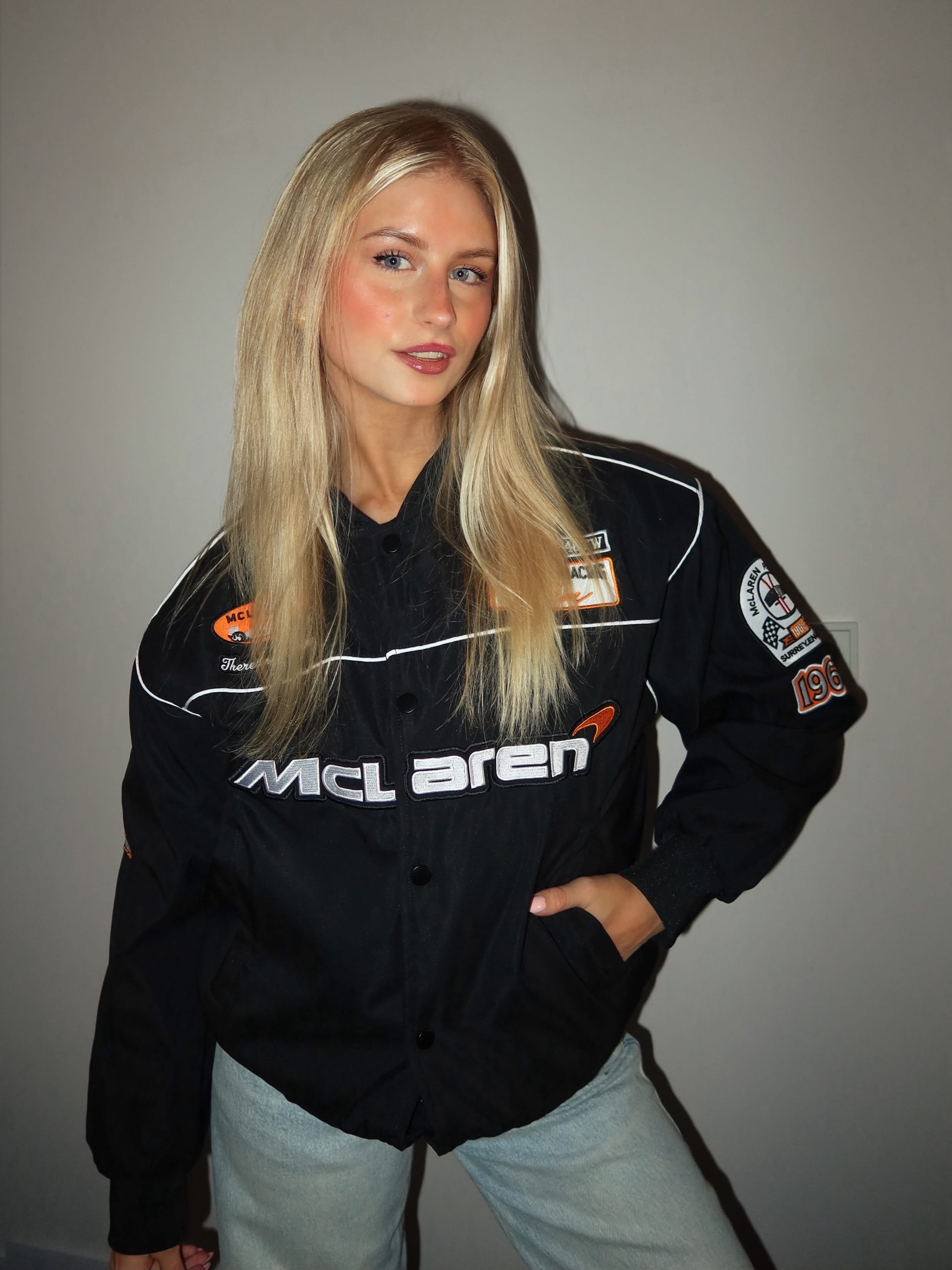 A woman with long blonde hair stands against a plain, light-colored wall, looking at the camera with a slight smile. She is wearing light blue jeans and the McLaren Vintage Bomber Jacket in black.