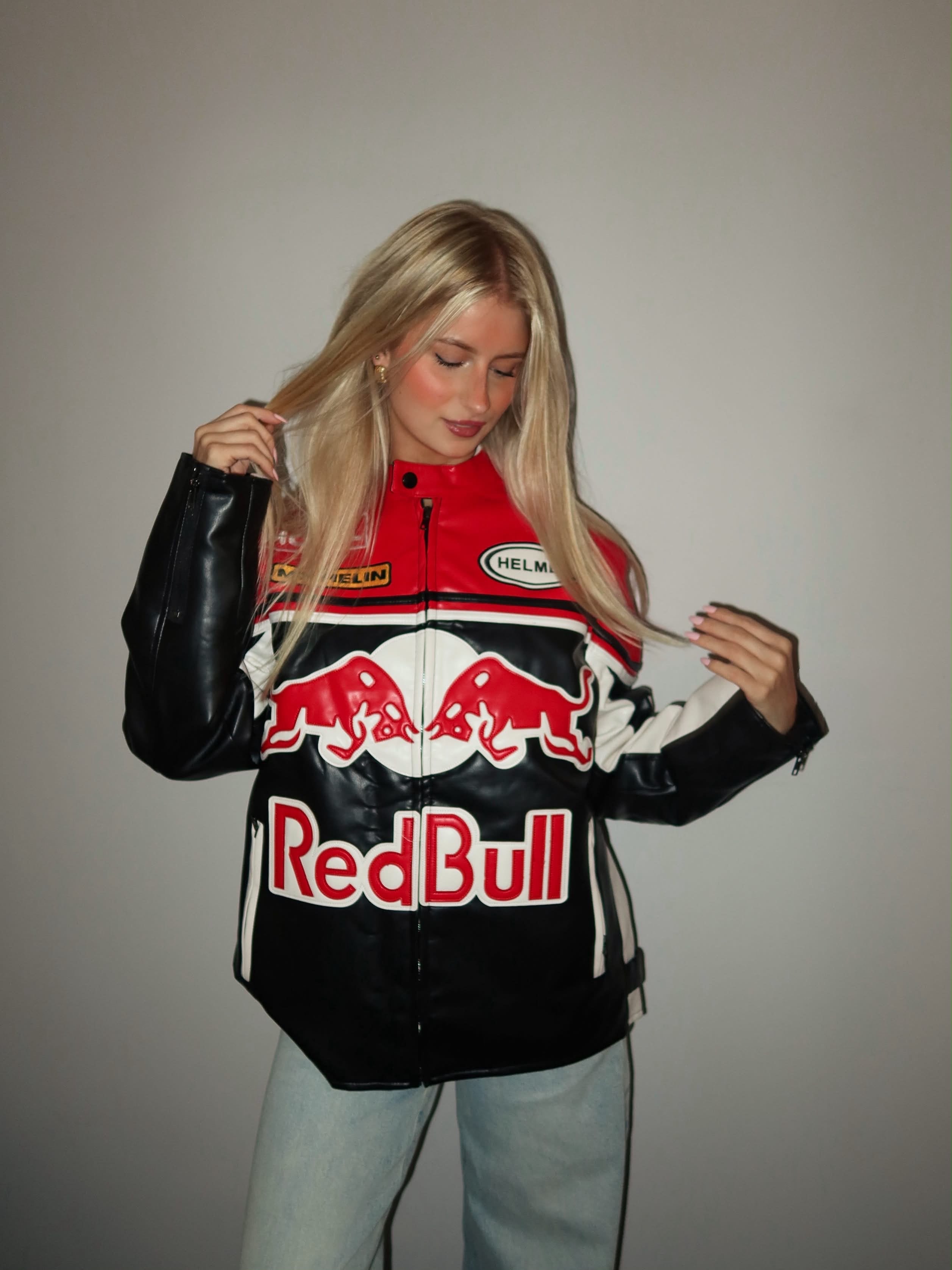 A woman with long blonde hair stands against a plain gray wall, looking down and touching her hair while wearing the Red Bull Leather Racing Jacket paired with light blue jeans.