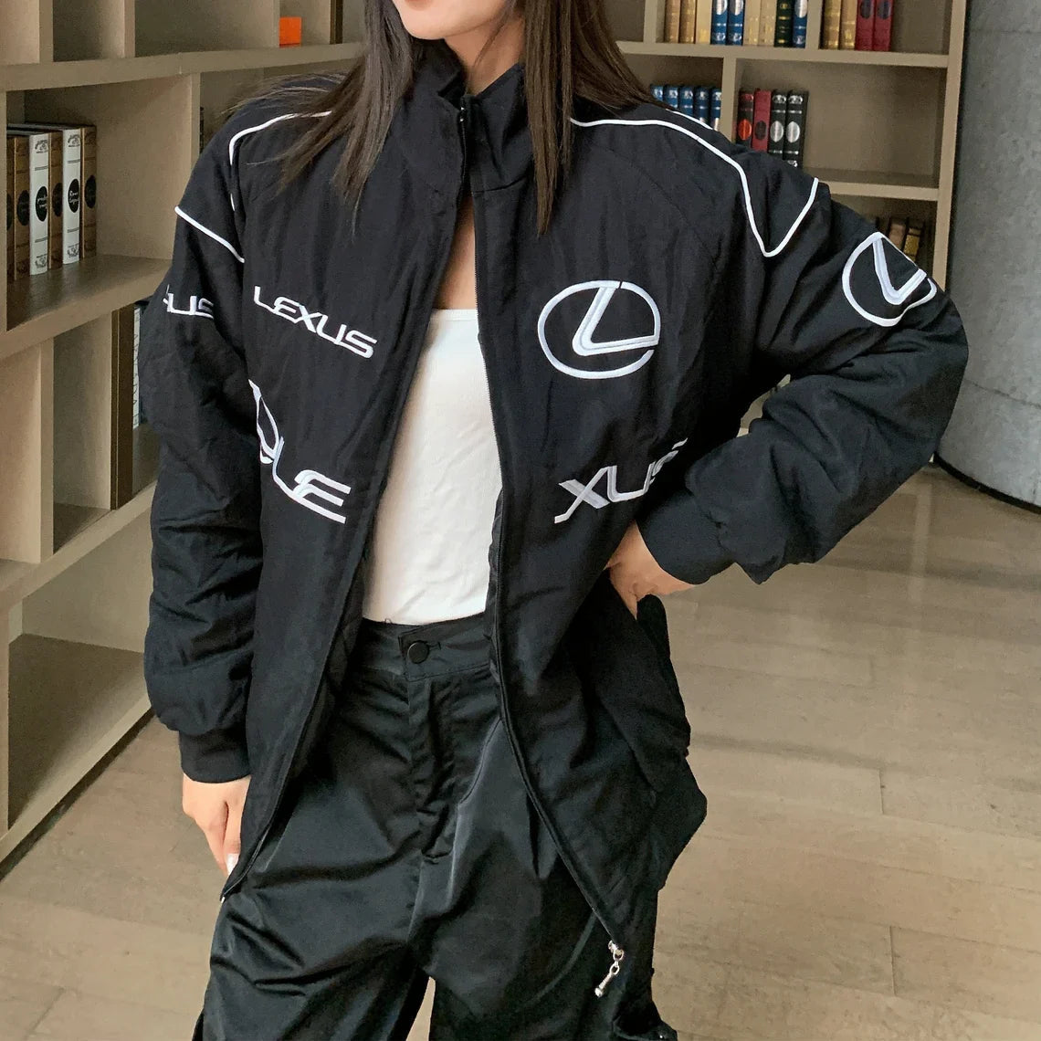 A woman stands indoors by bookshelves, wearing the Lexus Vintage Racing Jacket over a white top and black pants. Her face is partly out of frame, showcasing how this luxury motorsport piece can elevate everyday style.
