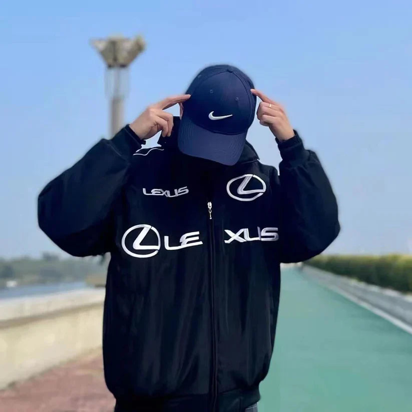 Wearing the Lexus Vintage Racing Jacket and a navy Nike cap, a person stands outdoors against a pathway and blue sky, head down and fingers pointing to the cap—showcasing luxury motorsport-inspired style.