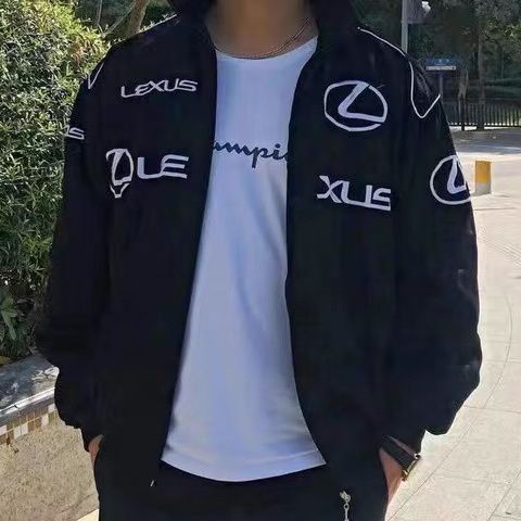Wearing the Lexus Vintage Racing Jacket, which features multiple white Lexus logos over a white T-shirt, a person stands outdoors with hands in pockets. The jacket channels luxury motorsport style; the wearers face is not shown.