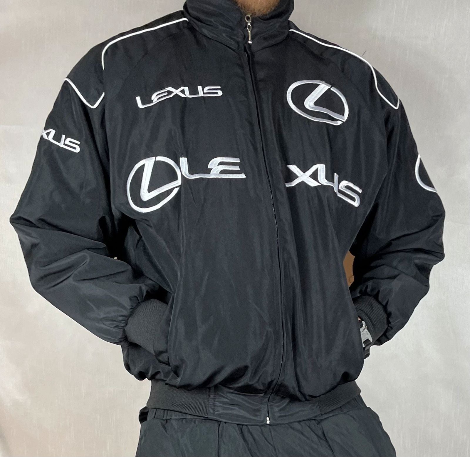 A person wears the Lexus Vintage Racing Jacket, a black motorsport-inspired piece with multiple white Lexus logos and bold LEXUS lettering on the chest, sleeves, and shoulders—a true statement of luxury racing style.