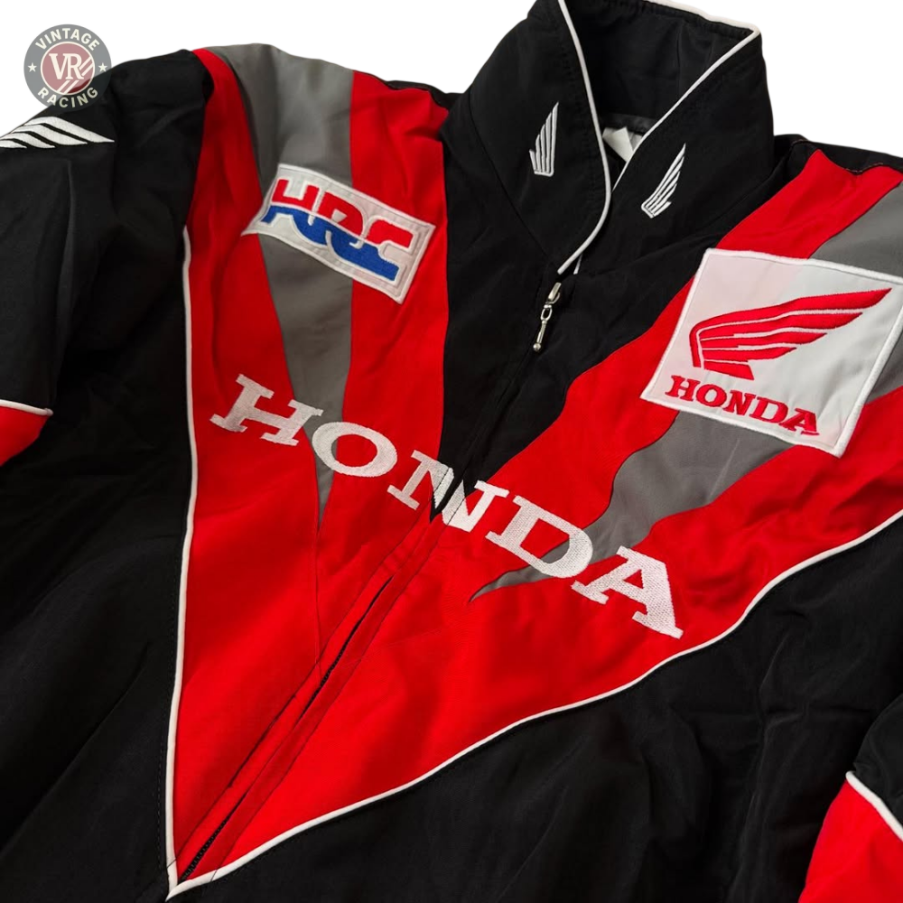 A close-up of the Honda Vintage Racing Jacket in black, red, and white with bold HONDA lettering, wing logos, and an HRC patch is displayed on a beige surface.