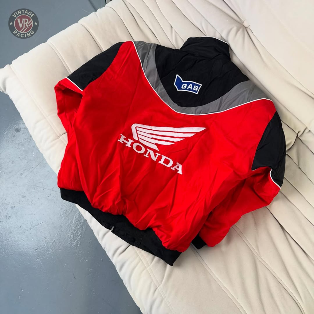 A Honda Vintage Racing Jacket featuring red, black, and gray colors with a bold Honda logo and GAS patch is showcased on a light-colored cushioned sofa.