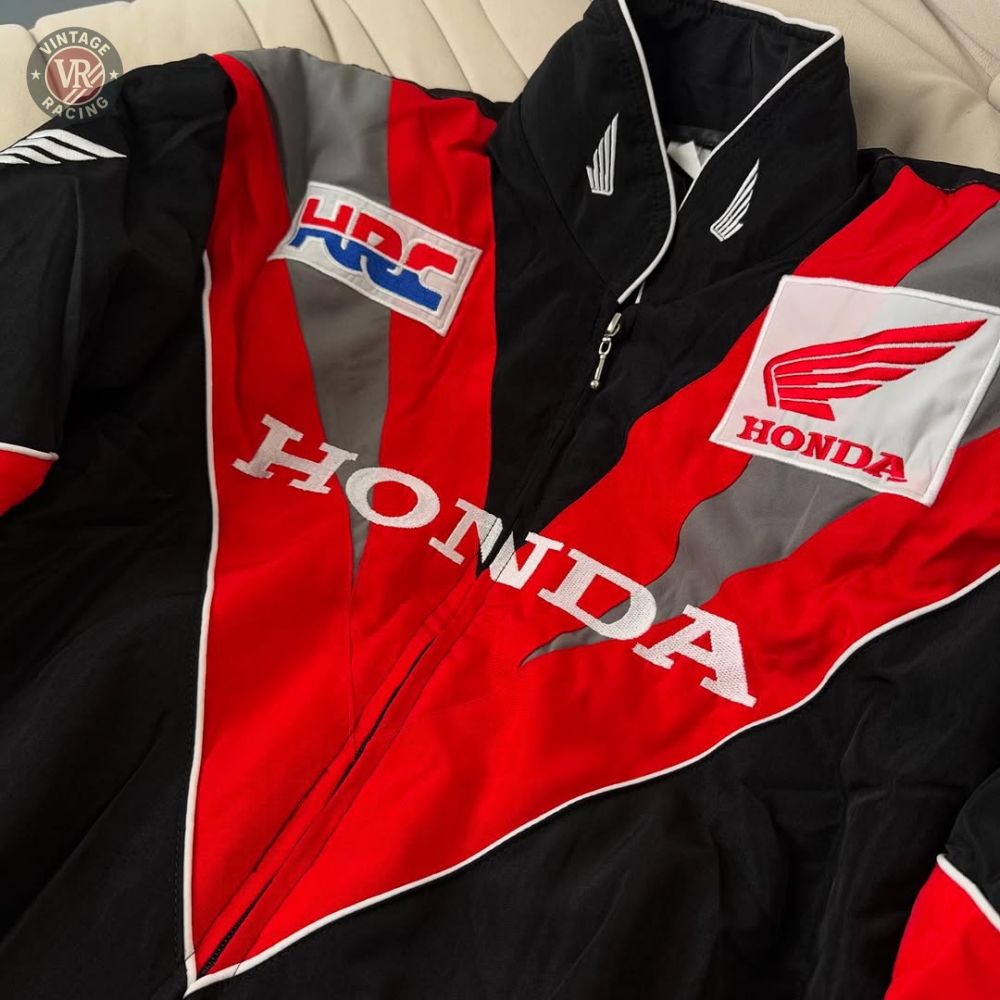 A close-up of the Honda Vintage Racing Jacket in black, red, and white with bold HONDA lettering, wing logos, and an HRC patch is displayed on a beige surface.