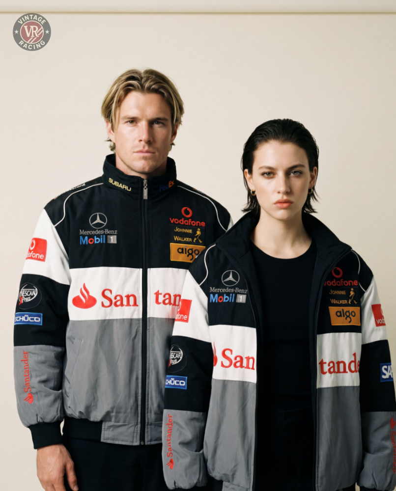 Two people stand side by side against a plain background, both wearing the McLaren F1 x Mercedes-Benz Vintage Jacket featuring black, gray, and white panels with sponsor logos. They face the camera with serious expressions.