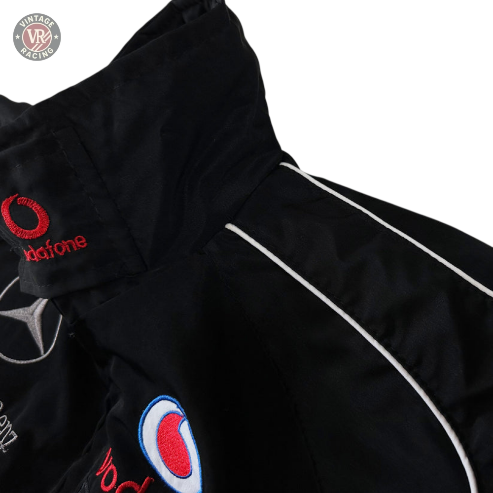 Close-up of the Mercedes-Benz Santander Vintage Jacket in black with white piping, featuring embroidered Vodafone, Mercedes star, and blue-red patches—highlighting its classic motorsport heritage on a light gray background.