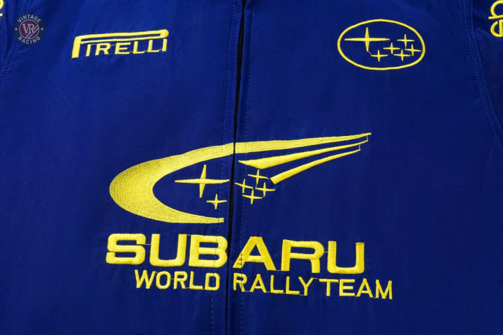 The Subaru World Rally Team Vintage Jacket is a blue fabric jacket with yellow embroidery featuring the team’s swoosh-and-stars logo, Pirelli branding, and Subaru logos inspired by the iconic 555 livery.
